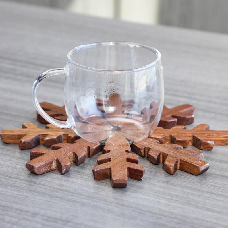 May include: A wooden snowflake coaster with a clear glass mug on top. The coaster is made of dark brown wood and has a detailed snowflake design.