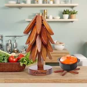 May include: A wooden kitchen utensil holder shaped like a tree, with a base that reads "Christine". An orange and black cooking pot sits on a wooden trivet. Fresh vegetables are in a basket.