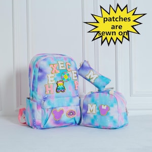 May include: A blue and pink tie-dye backpack set with a lunch bag, pencil case, and a backpack. The backpack has patches sewn on it, including letters, a bear, a donut, and a Mickey Mouse head.