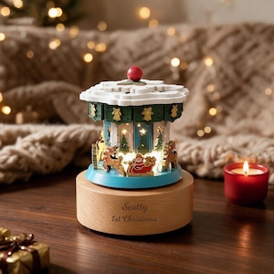 May include: A wooden carousel music box featuring a white snowflake roof with a red knob, green panels with gold bell accents, and illuminated scenes of Santa in a sleigh with reindeer. The base is light wood engraved with Scotty 1st Christmas.