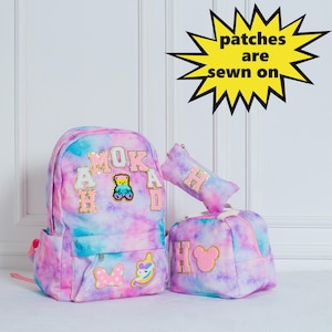 May include: A tie-dye backpack, pencil case, and small tote bag set. The bags are in shades of pink, blue, and purple. The backpack has the letters "MOKAD" and a teddy bear patch. The pencil case and tote bag have the letter "H".