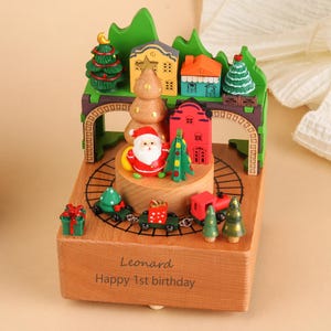 May include: A wooden Christmas music box featuring a Santa Claus figure, miniature houses, and a train. The box has a green arch and a wooden base with the text "Leonard Happy 1st birthday".