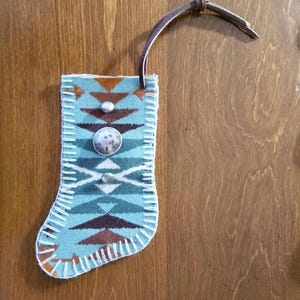 May include: A small, turquoise Christmas stocking with a geometric pattern in brown and white. The stocking is decorated with two silver-colored buttons and a brown leather loop for hanging. The stocking is made of a felt-like material.