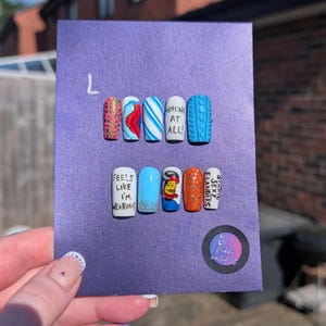Puede incluir: Un conjunto de uñas postizas sobre un fondo morado. Las uñas presentan varios diseños, incluyendo patrones, texto y personajes de dibujos animados. El texto en las uñas incluye frases como "NOTHING AT ALL!" y "FEELS LIKE I'M WEARING".