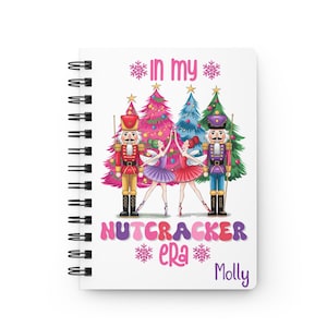 Peut inclure: Un carnet à spirales blanc avec un motif d'arbre de Noël rose, bleu et vert. Le texte "In my Nutcracker era" est imprimé en lettres roses et blanches. Le nom "Molly" est imprimé en lettres violettes en bas du carnet.