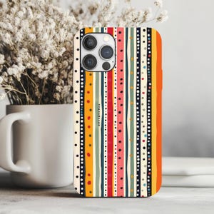 May include: A colorful phone case with a vertical striped pattern. The design features stripes in orange, yellow, red, green, and blue, interspersed with dotted lines. The case is shown next to a white mug and a vase of dried flowers.