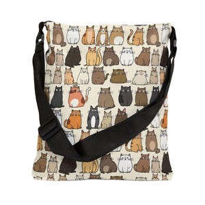 May include: A beige canvas tote bag with a black adjustable shoulder strap. The bag features a repeating pattern of colorful cartoon cats in various poses.