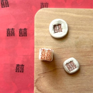 May include: Three white clay stamps with the Chinese character for "double happiness" in red ink. The stamps are on a wooden surface.