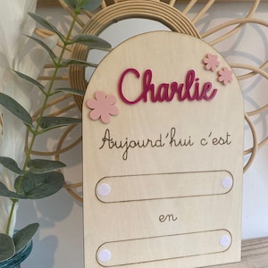 Peut inclure: Un panneau en bois clair avec le prénom "Charlie" en rose, orné de fleurs roses. Le panneau comprend le texte français "Aujourd'hui c'est" et "en", avec deux emplacements pour la personnalisation.