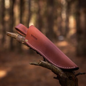 Könnte beinhalten: Eine braune Ledertasche für ein Messer mit weißen Nähten und einem Messingverschluss. Die Tasche hat den Text "GPSKNIVES" auf der Vorderseite geprägt.