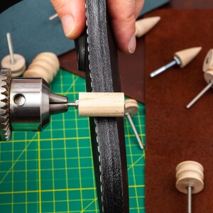 Puede incluir: Primer plano de un proyecto de artesan&iacute;a en cuero que se est&aacute; trabajando con un taladro y una clavija de madera. El cuero es negro y tiene costuras alrededor del borde. La clavija se est&aacute; insertando en el cuero con el taladro.