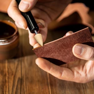 Puede incluir: Una persona usando una herramienta de trabajo de cuero con una punta de madera para crear un pliegue en un trozo de cuero marr&oacute;n.