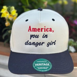 Op de afbeelding: Crèmekleurige baseballcap met een marineblauwe klep. De cap heeft de tekst "America, you in danger girl" in rode en zwarte letters. Een groene en witte "Legacy Heritage" sticker staat op de klep.