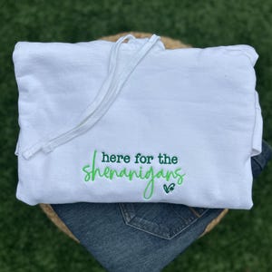 May include: White hoodie with a drawstring, featuring the embroidered phrase "here for the shenanigans" in green script. The hoodie is folded over a pair of blue jeans, set against a green background.