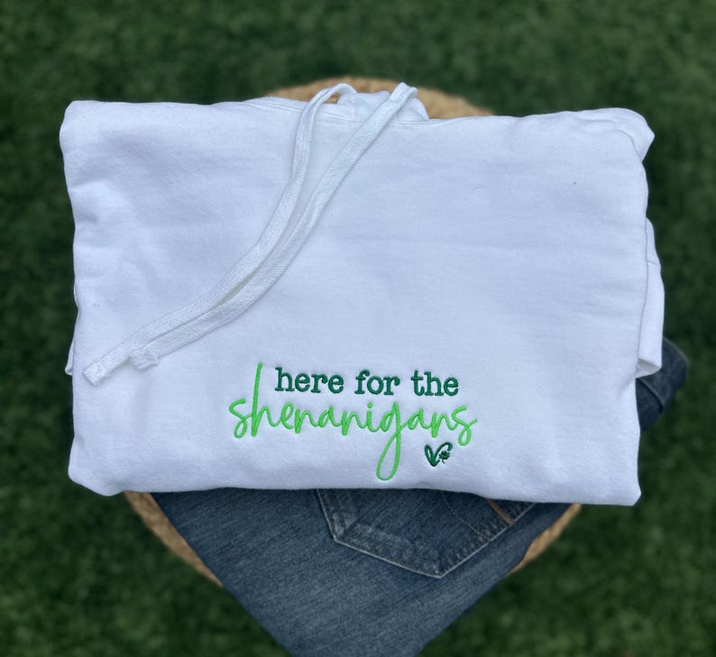 May include: White hoodie with a drawstring, featuring the embroidered phrase "here for the shenanigans" in green script. The hoodie is folded over a pair of blue jeans, set against a green background.