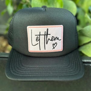 May include: Black trucker hat with a mesh back and a curved brim. The front features a rectangular patch with the words "Let them" in black script, and a small heart. The patch has a pink border.