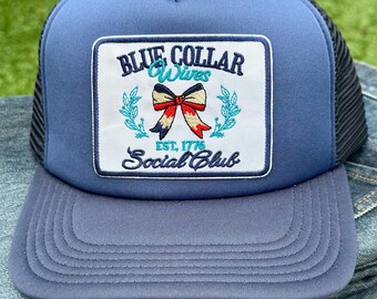 Può includere: Un cappello da camionista blu con una rete nera sul retro. La parte anteriore del cappello presenta una toppa bianca con un fiocco rosso, bianco e blu e il testo "Blue Collar Wives Est. 1776 Social Club".