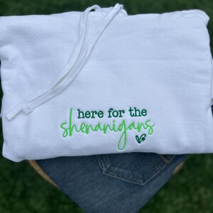 May include: A white hooded sweatshirt with a drawstring, featuring the embroidered phrase "I'm here for the shenanigans" in green script. The sweatshirt is folded over a pair of blue jeans.