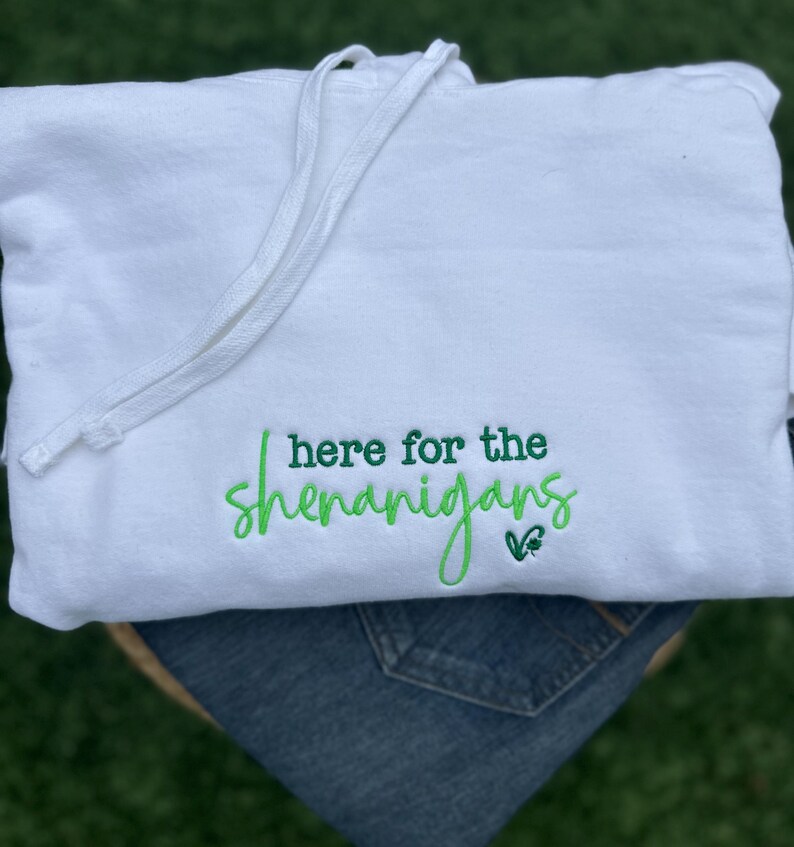 May include: A white hooded sweatshirt with a drawstring, featuring the embroidered phrase "I'm here for the shenanigans" in green script. The sweatshirt is folded over a pair of blue jeans.