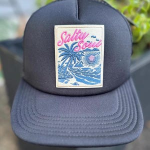 May include: A black trucker cap featuring a rectangular patch on the front. The patch displays an embroidered coastal scene with a palm tree, ocean waves, and a sun, with the text Salty Soul stitched in pink script lettering above the graphic.