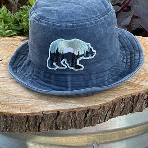 May include: A blue denim bucket hat with a patch of a bear silhouette in front of a mountain range with a rainbow.