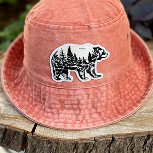 May include: A coral-coloured bucket hat with a black and white embroidered patch of a bear with trees inside its silhouette.
