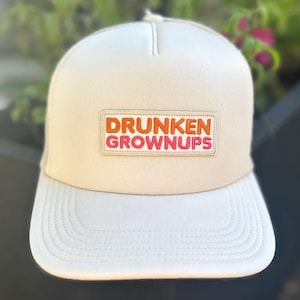 May include: A cream-colored baseball cap featuring a rectangular patch on the front. The patch displays the text "DRUNKEN" in orange embroidery on the top line and "GROWNUP" in pink embroidery on the bottom line, all on a light background.