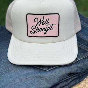 Op de afbeelding: Een witte truckercap met een roze patch waarop in zwarte letters "Well Sheeyit" staat.