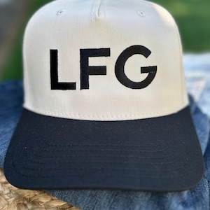 May include: A beige and black baseball cap featuring the embroidered letters "LFG" in black. The cap has a structured front panel and a curved brim. The hat is made of a durable fabric.
