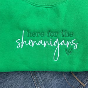 May include: A bright green sweatshirt with the embroidered phrase "here for the shenanigans" in white script. The sweatshirt is folded over a pair of blue jeans. A small green clover is embroidered next to the word "shenanigans".