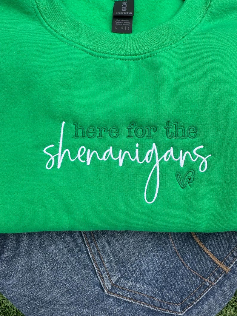 May include: A bright green sweatshirt with the embroidered phrase "here for the shenanigans" in white script. The sweatshirt is folded over a pair of blue jeans. A small green clover is embroidered next to the word "shenanigans".
