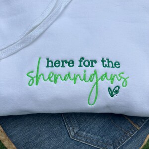 May include: White hooded sweatshirt with the embroidered phrase "here for the shenanigans" in green script. A small green shamrock is next to the text. The sweatshirt is paired with blue denim.