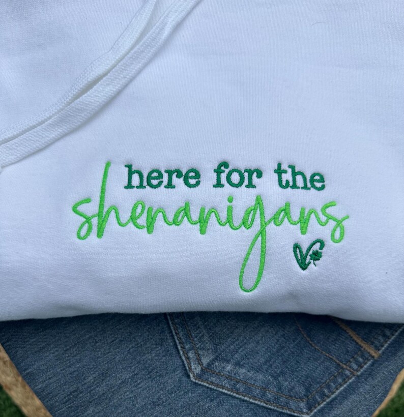 May include: White hooded sweatshirt with the embroidered phrase "here for the shenanigans" in green script. A small green shamrock is next to the text. The sweatshirt is paired with blue denim.