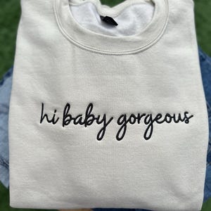 Puede incluir: Una sudadera gris claro con el texto "hi baby gorgeous" bordado en negro.