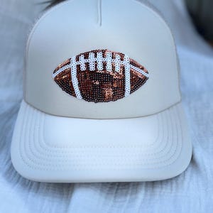 Könnte beinhalten: Ein weißer Trucker-Hut mit einem braunen und weißen Football-Design aus Pailletten auf der Vorderseite.
