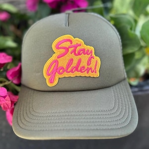 Puede incluir: Gorra de camionero verde oliva con visera curva y parte trasera de malla. La gorra presenta un parche con las palabras "Stay Golden!" en rosa y amarillo. Un accesorio casual.