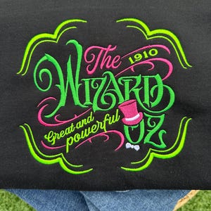 Op de afbeelding: Een groen en roze geborduurd ontwerp op een zwart T-shirt met de tekst "The Wizard of Oz" met het jaar "1910" boven het woord "Wizard" en de tekst "Great and powerful" onder het woord "Wizard".