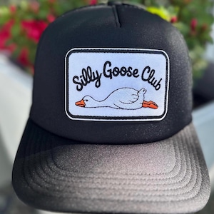 Puede incluir: Gorra de camionero negra con un parche blanco con el texto "Silly Goose Club" y un dibujo de un ganso. El ganso es blanco con pico y patas naranjas. La gorra tiene una visera negra y una parte trasera de malla.