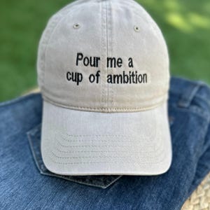 May include: A beige baseball cap with black embroidered text that reads "Pour me a cup of ambition".