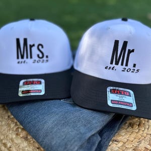 Puede incluir: Dos gorras de béisbol blancas y negras con las palabras "Mrs. est. 2025" y "Mr. est. 2025" bordadas.
