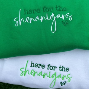 May include: Two sweatshirts, one green and one white, are displayed. The green sweatshirt has the phrase "here for the shenanigans" embroidered in white script. The white sweatshirt has the same phrase embroidered in green script.