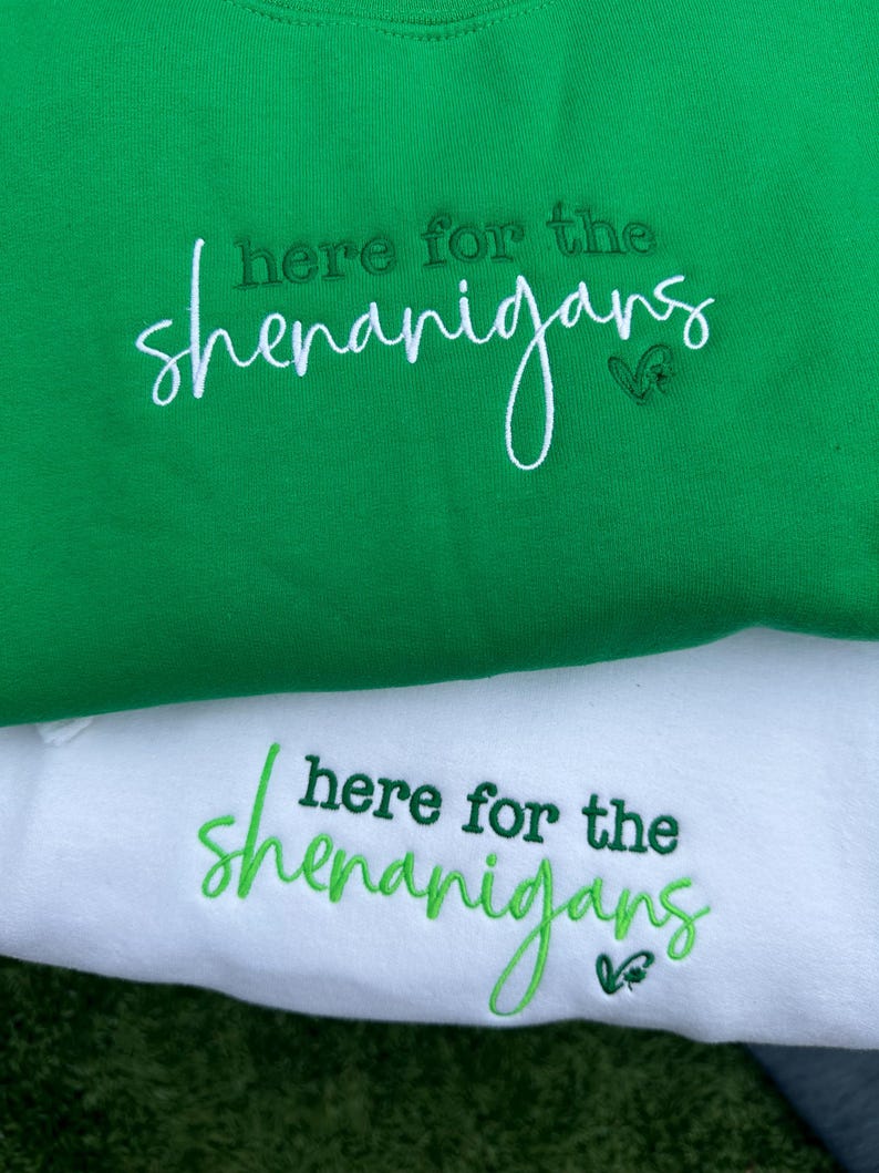 May include: Two sweatshirts, one green and one white, are displayed. The green sweatshirt has the phrase "here for the shenanigans" embroidered in white script. The white sweatshirt has the same phrase embroidered in green script.