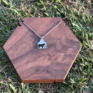 May include: A silver necklace with a pendant shaped like a cow tag. The tag is white with a black cow silhouette and the letter "R". The necklace is displayed on a brown wooden hexagon.