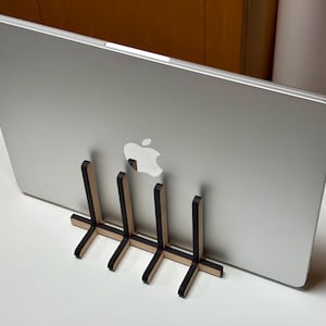 Peut inclure: Un ordinateur portable argenté avec le logo Apple est maintenu par un support en bois. Le support a une base en forme de croix et des supports verticaux. L'ordinateur portable est posé sur une surface blanche.