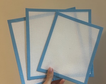 May include: Three rectangular needlepoint canvases with a white mesh and a light blue border. The canvases are held in a hand against a neutral background. The mesh is tightly woven with small holes.