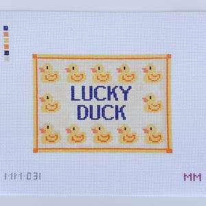 May include: Needlepoint canvas featuring a grid of yellow ducklings surrounding the words "LUCKY DUCK" in blue. The design is framed in orange on a white background. The canvas has a grid-like texture.