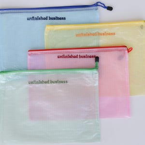 May include: A collection of colorful mesh zipper pouches in blue, yellow, pink, and green. Each pouch has the words "unfinished business" printed on it. The pouches are designed for organizing and storing items.