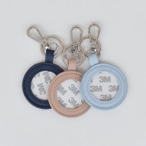 May include: Three circular keychains in navy blue, dusty pink, and light blue. Each features a silver metal lobster clasp and a central white adhesive backing with the repeating text 3M printed in grey across the surface of the circular centre.