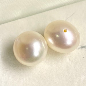 May include: Two white round pearls with a hole drilled through the center. The pearls are sitting on a white surface.