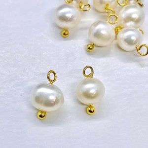 May include: A close-up of two white pearl charms with gold jump rings and a gold bead at the bottom. The charms are on a white surface.